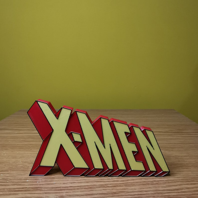 K&ouml;nnte beinhalten: Ein rotes und gelbes 3D-Plastikschild mit der Aufschrift "X-Men".