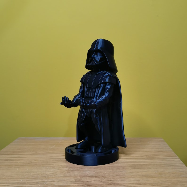May include: A black 3D printed figurine of Darth Vader from Star Wars. The figurine is standing on a round base and has a cape.