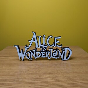 Pode incluir: Uma placa de madeira branca e preta que diz "Alice in Wonderland". A placa &eacute; colocada em uma superf&iacute;cie de madeira com um fundo amarelo. A escrita &eacute; em uma fonte caprichosa.