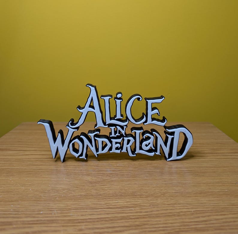 Pode incluir: Uma placa de madeira branca e preta que diz "Alice in Wonderland". A placa &eacute; colocada em uma superf&iacute;cie de madeira com um fundo amarelo. A escrita &eacute; em uma fonte caprichosa.