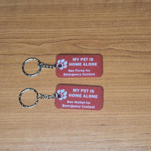 May include: Two red keychains with silver key rings. The keychains have white paw prints and the text "MY PET IS HOME ALONE" with instructions to see the purse or wallet for emergency contact.