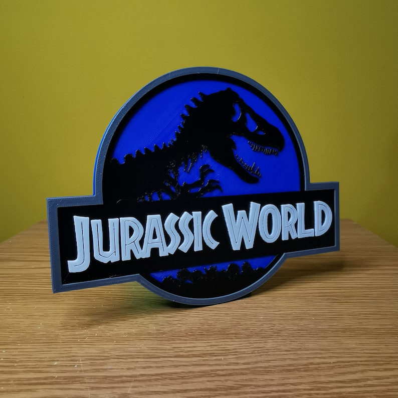 May include: A 3D printed Jurassic World logo with a blue background and a black silhouette of a dinosaur. The text "Jurassic World" is in white.