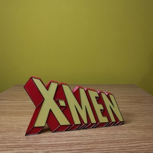 K&ouml;nnte beinhalten: Ein 3D-gedrucktes Schild mit der Aufschrift "X-Men" in roten und gelben Buchstaben mit schwarzen Umrissen.