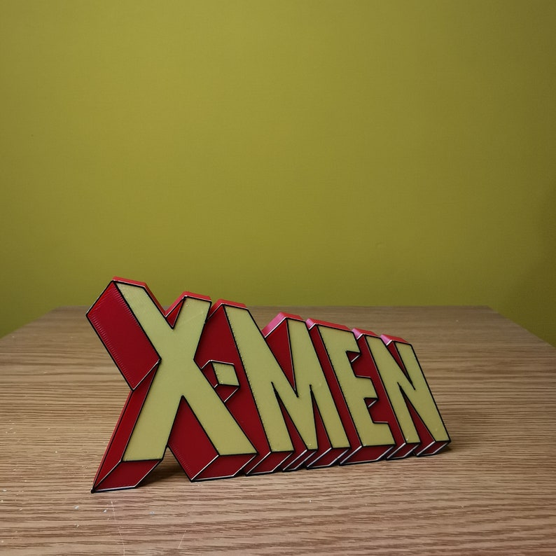 K&ouml;nnte beinhalten: Ein 3D-gedrucktes Schild mit der Aufschrift "X-Men" in roten und gelben Buchstaben mit schwarzen Umrissen.