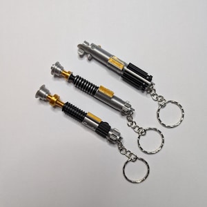 May include: Three silver lightsaber keychains with black, gold, and silver detailing. Each keychain has a silver ring attached. The lightsabers are arranged diagonally on a white surface.