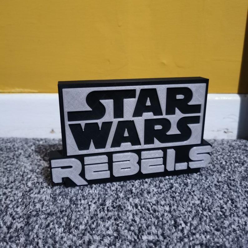 May include: Black and silver 3D printed sign with the text "STAR WARS REBELS".