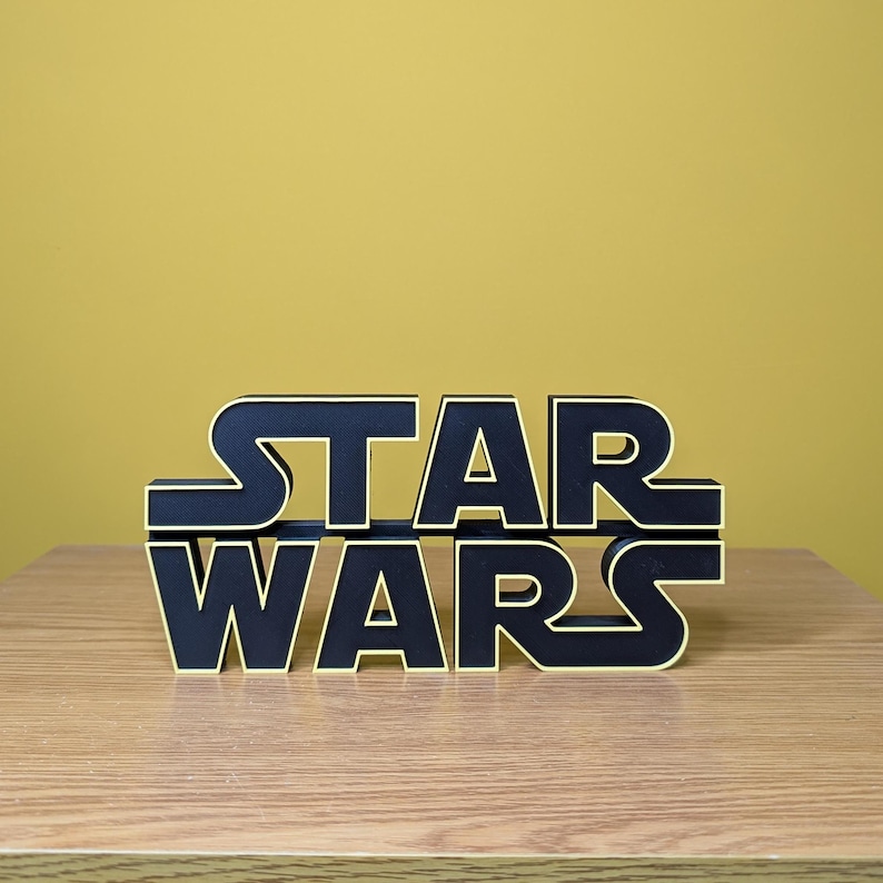 Peut inclure: Un panneau du logo Star Wars noir et or. Les lettres sont noires avec un contour dor&eacute;, et le panneau est affich&eacute; sur une surface en bois sur un fond jaune. Le panneau est un objet d&eacute;coratif.