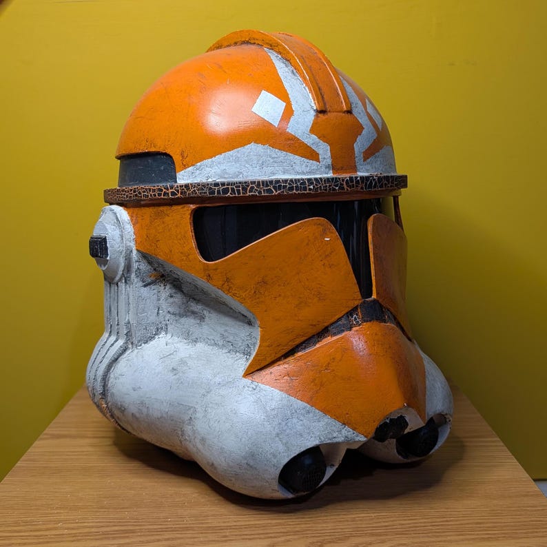 May include: A white and orange plastic Star Wars clone trooper helmet with a weathered finish. The helmet has a white visor and a white stripe across the top.