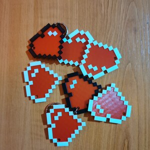 May include: Six red pixel heart-shaped keychains with a light blue border. The keychains are made of plastic and have a black metal ring.