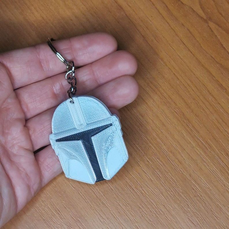 May include: A white and black 3D printed keychain featuring a Mandalorian helmet design. The keychain has a silver metal ring.