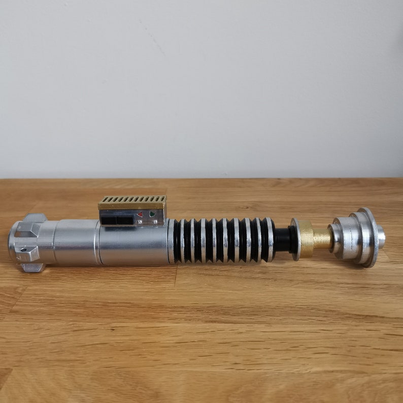 May include: A silver and black lightsaber prop with a gold-colored handle and a black ribbed section. The lightsaber has a small, rectangular, silver control panel with a green and red light.
