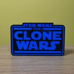 以下が含まれることがあります： 白の四角形の中に「Star Wars The Clone Wars」という文字が書かれた、黒と青の3Dプリントされた看板です。