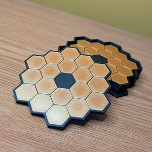 Puede incluir: Un juego de cuatro posavasos hexagonales negros y dorados. Los posavasos están hechos de un material impreso en 3D y tienen un patrón de panal.