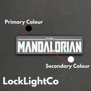 May include: A black and white graphic of "The Mandalorian" logo. The logo is on a grey background. The primary colour is black and the secondary colour is white.