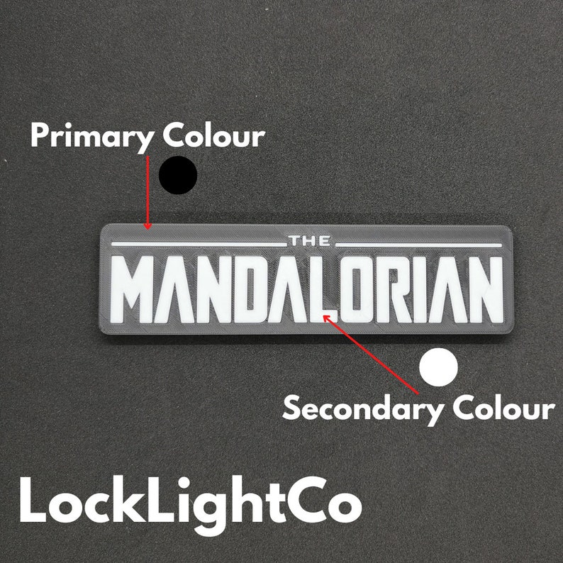 May include: A black and white graphic of "The Mandalorian" logo. The logo is on a grey background. The primary colour is black and the secondary colour is white.