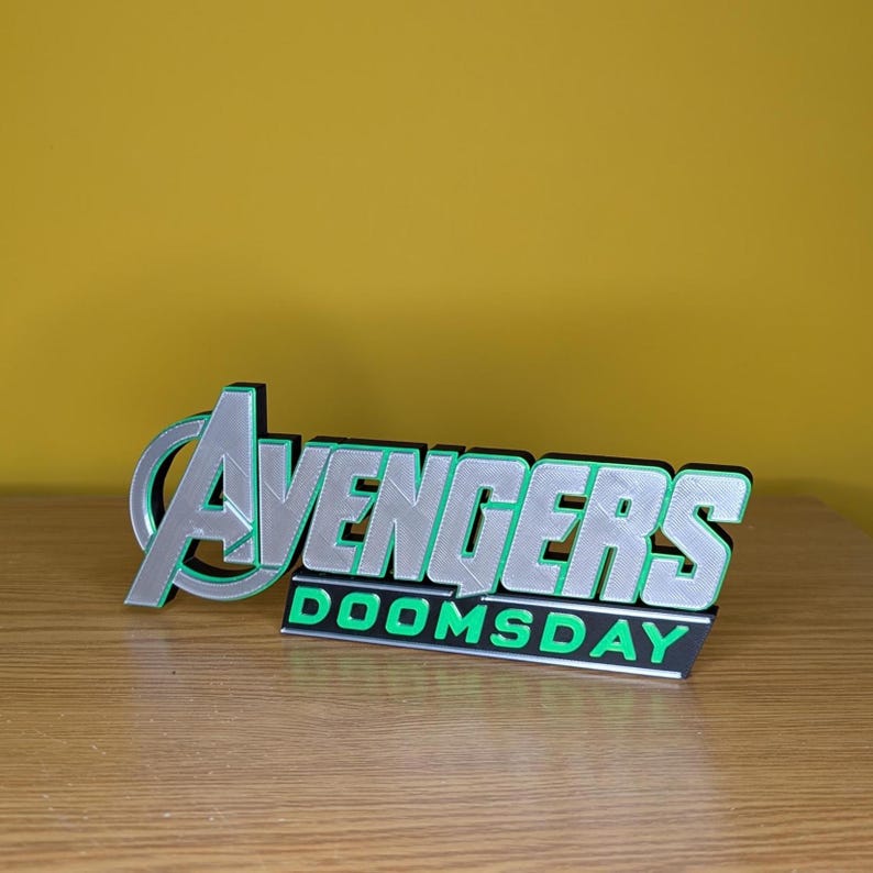 以下が含まれることがあります： 3Dプリントされた、シルバーとグリーンの「Avengers Doomsday」ロゴ。緑色の輪郭を持つロゴは、スタイリッシュなフォントで「Avengers」の文字と、その下に「Doomsday」の文字を表示しています。木製の表面に置かれています。