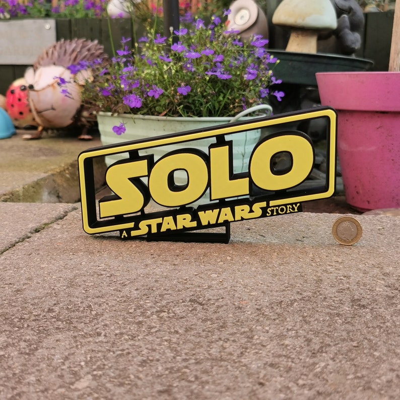 May include: A black and yellow sign with the words "Solo: A Star Wars Story" in a bold, stylized font. The sign is sitting on a gray concrete surface.
