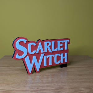 May include: A Scarlet Witch sign with a red background and white lettering. The sign has a black border and is standing on a wooden surface. The background is a solid yellow color.