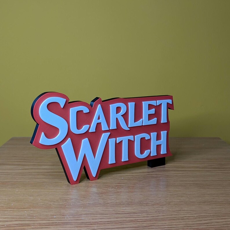 May include: A Scarlet Witch sign with a red background and white lettering. The sign has a black border and is standing on a wooden surface. The background is a solid yellow color.