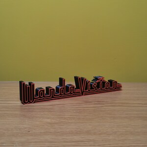Peut inclure: Un panneau imprim&eacute; en 3D avec le texte "WandaVision". Les lettres noires sont bord&eacute;es de rouge. Le panneau est pos&eacute; sur une surface en bois, devant un fond jaune.