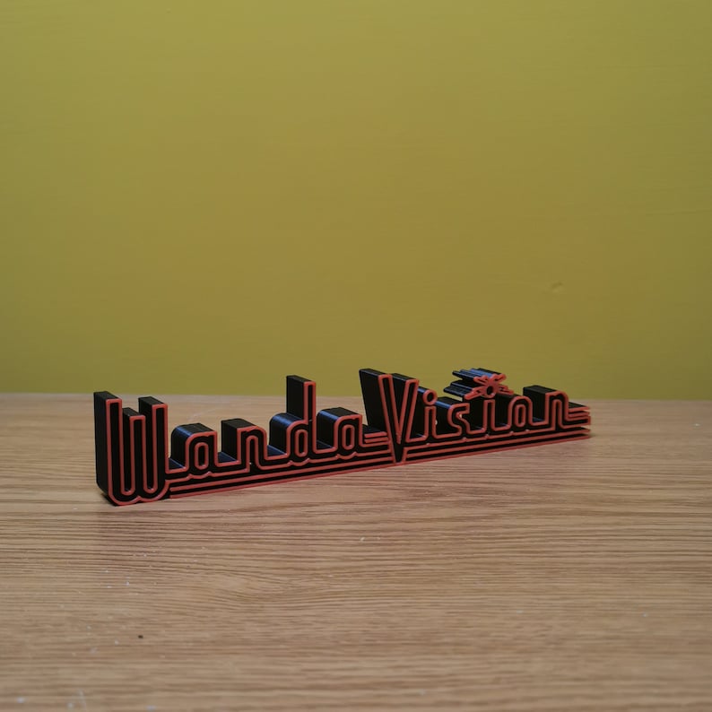 Peut inclure: Un panneau imprim&eacute; en 3D avec le texte "WandaVision". Les lettres noires sont bord&eacute;es de rouge. Le panneau est pos&eacute; sur une surface en bois, devant un fond jaune.