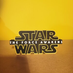 May include: Black 3D printed Star Wars logo with yellow outline. The text "The Force Awakens" is printed below the logo.