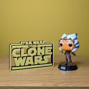 May include: A yellow and black 3D printed sign that reads "Star Wars The Clone Wars". A small figurine of Ahsoka Tano from Star Wars The Clone Wars is standing on a black base.