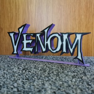 May include: A 3D-printed sign displaying the word "VENOM". The letters are white with black edging, set against a purple backing. The sign is designed to stand upright. The overall design is a stylized, three-dimensional representation.