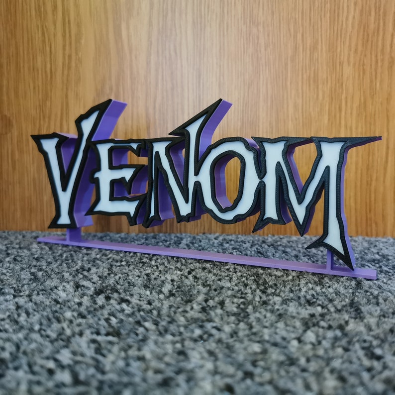 May include: A 3D-printed sign displaying the word "VENOM". The letters are white with black edging, set against a purple backing. The sign is designed to stand upright. The overall design is a stylized, three-dimensional representation.