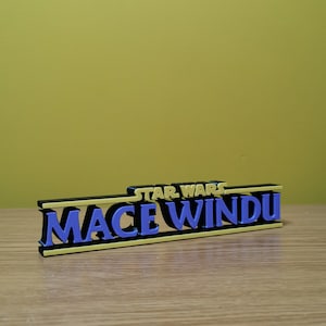 Peut inclure: Une enseigne en 3D imprimée en noir et or avec le texte "STAR WARS MACE WINDU" en lettres violettes.