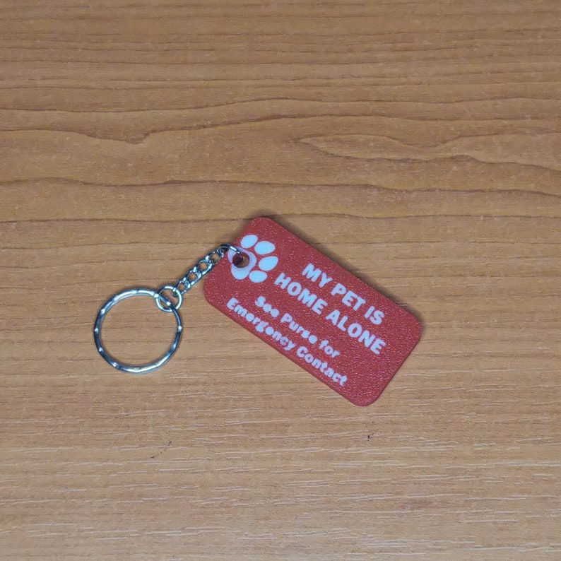 May include: Red rectangular keychain with white text that reads "My Pet is Home Alone. See Purse for Emergency Contact". The keychain has a silver ring and chain.