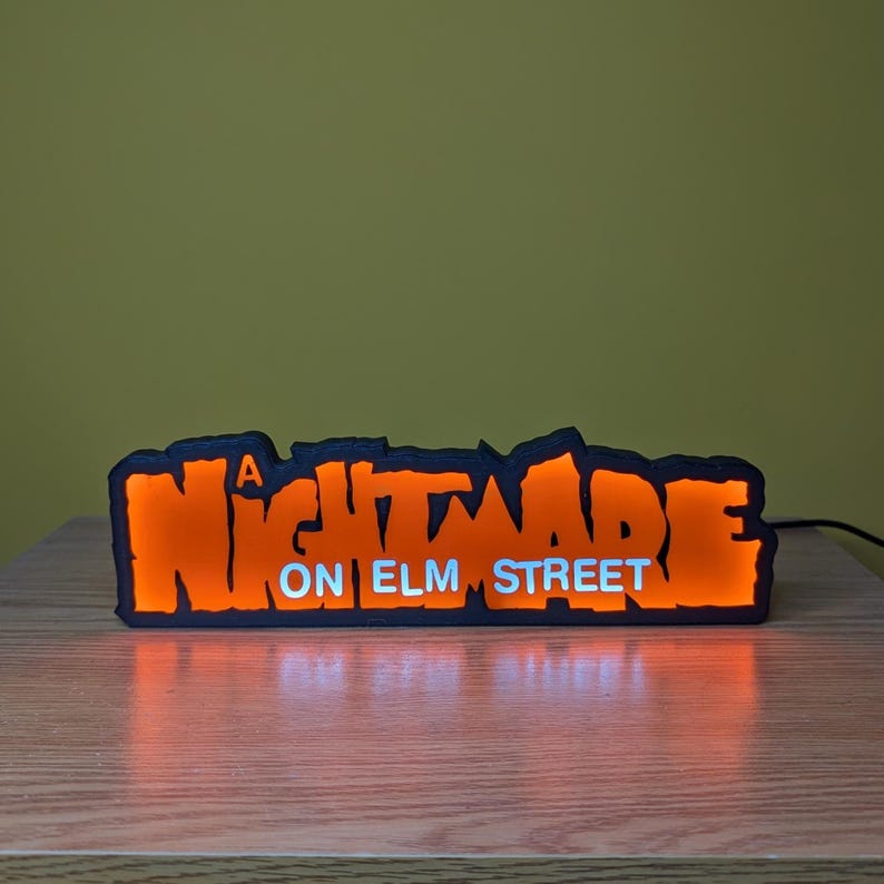 May include: A black and orange LED sign with the text "A NIGHTMARE ON ELM STREET". The sign is illuminated with orange light, and the text is in a stylized font. The sign is on a wooden surface.