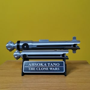 May include: A silver lightsaber prop with a black handle and a silver stand. The stand has a black plaque with the text "Ahsoka Tano The Clone Wars".