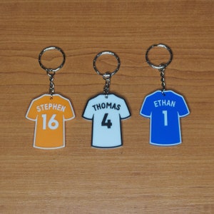 Puede incluir: Tres llaveros con forma de camiseta. Uno es naranja con "STEPHEN" y el n&uacute;mero "16", otro es blanco con "THOMAS" y el n&uacute;mero "4", y el &uacute;ltimo es azul con "ETHAN" y el n&uacute;mero "1". Cada llavero tiene un aro plateado.