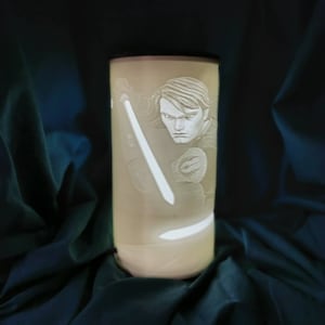 May include: A white ceramic lamp with a 3D design of a character holding a lightsaber. The character has short, dark hair and is wearing a dark outfit.