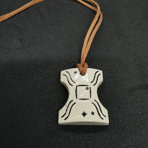May include: A white ceramic pendant with a geometric design and black lines. The pendant is attached to a brown leather cord.
