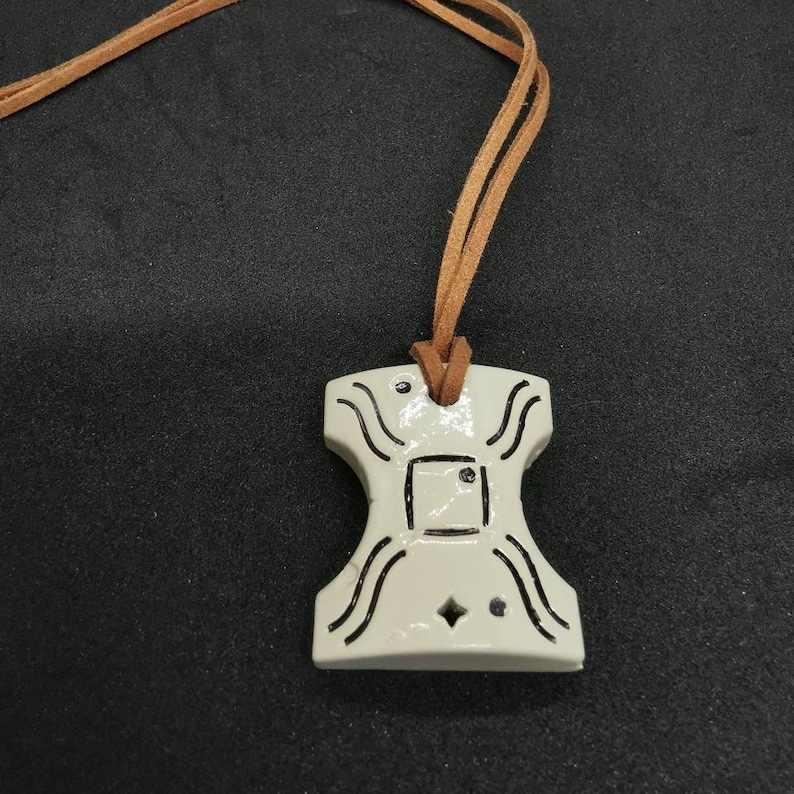 May include: A white ceramic pendant with a geometric design and black lines. The pendant is attached to a brown leather cord.