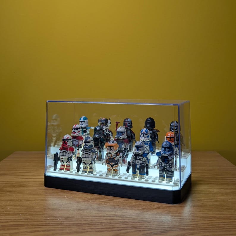May include: A clear plastic display case with a black base, showcasing a collection of Star Wars Lego minifigures. The minifigures are arranged in a row, with a variety of characters from the Clone Wars era. The case is illuminated from below, highlighting the details of the figures.