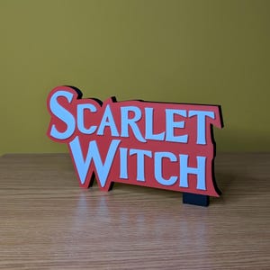 May include: A red and white Scarlet Witch sign with a black border. The sign is made of layered material and stands upright on a small black base. The text is in a bold, stylized font.