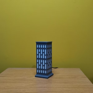 May include: A gray table lamp shaped like a building with a white light inside. The lamp is on a wooden table.