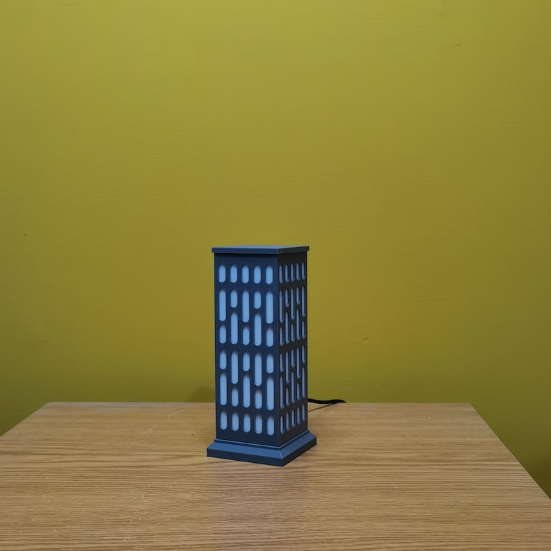 May include: A gray table lamp shaped like a building with a white light inside. The lamp is on a wooden table.