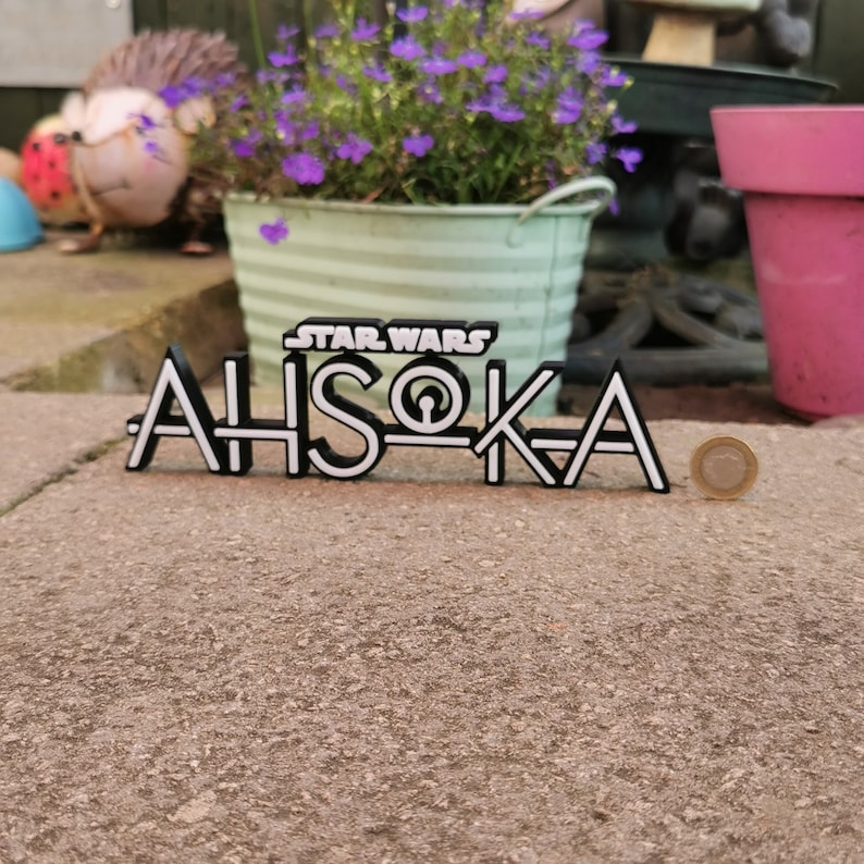 May include: A black and white Star Wars "Ahsoka" sign. The sign is made of layered material, with the text "STAR WARS" above "AHSOKA". A coin is next to the sign. The sign is on a concrete surface, with a flower pot in the background.