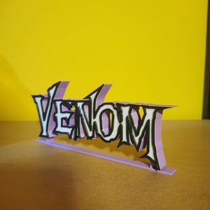 May include: A 3D-printed sign featuring the word "Venom" in a distinctive font. The letters are white with black borders, set against a light purple base. The sign is designed to stand on a flat surface.