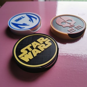 May include: Three round coasters with different designs. The first coaster is white with a blue and red design and the text "301". The second coaster is yellow with a red skull and crossbones design and the text "555". The third coaster is black with a yellow "STAR WARS" logo.