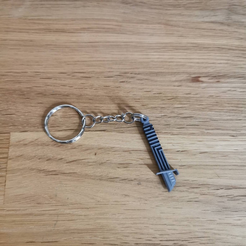 Op de afbeelding: Een zilveren sleutelhanger met een zilveren ring en een zilveren ketting bevestigd aan een grijze plastic zwaardbedel.