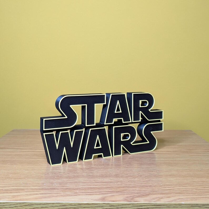 Peut inclure: Logo "Star Wars" en noir et jaune, imprim&eacute; en 3D, pos&eacute; sur une surface en bois. Les lettres noires sont bord&eacute;es de jaune, sur un fond jaune uni. Objet d&eacute;coratif.