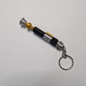 May include: A miniature lightsaber keychain with a silver, black, and gold design. The keychain features a detailed replica of a lightsaber, complete with a ring for attaching keys. The lightsaber is approximately 5 inches long.