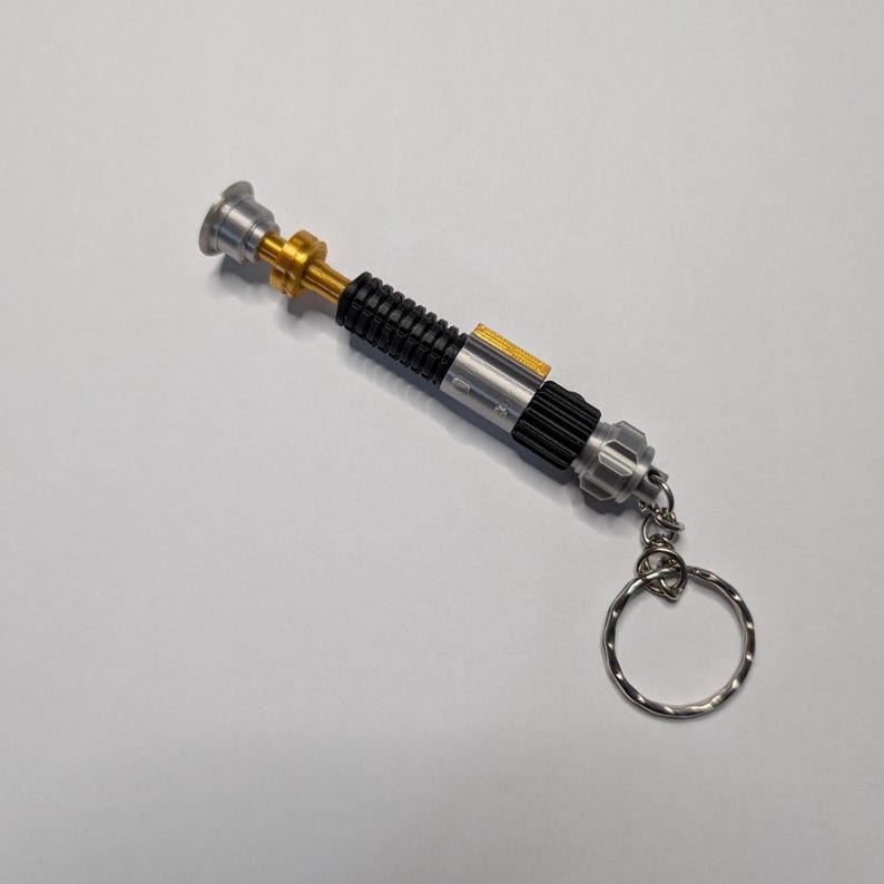 May include: A miniature lightsaber keychain with a silver, black, and gold design. The keychain features a detailed replica of a lightsaber, complete with a ring for attaching keys. The lightsaber is approximately 5 inches long.