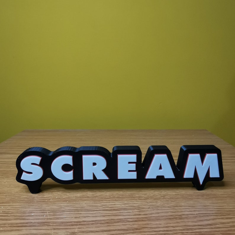 May include: Black and white 3D word sign that reads "SCREAM" with red outlines. The sign is on a wooden surface.