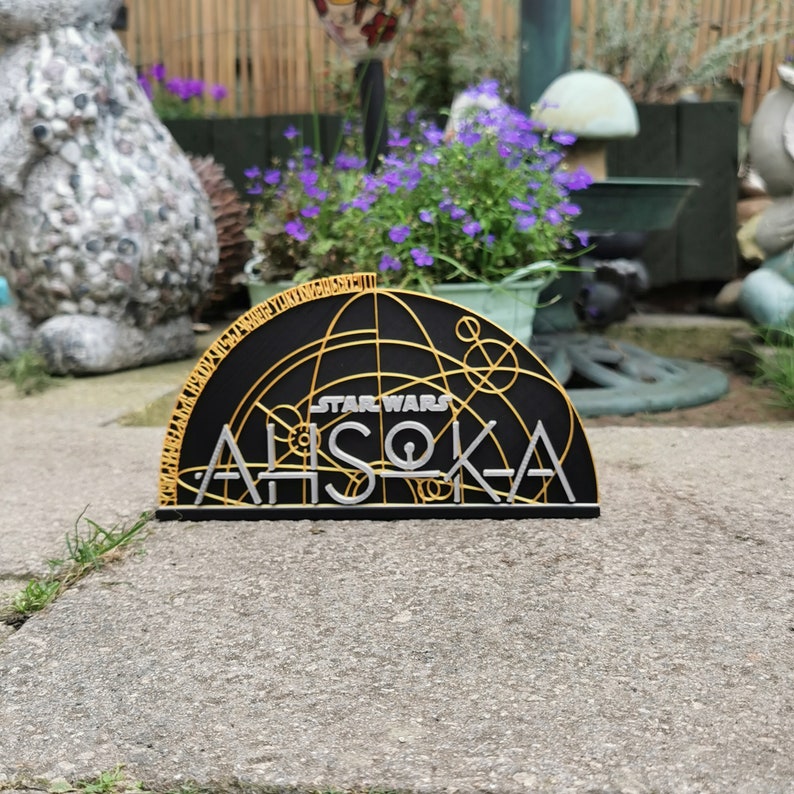 Może przedstawiać: Czarno-złote logo Star Wars Ahsoka Tano z okrągłym wzorem. Logo znajduje się na czarnym tle ze złotymi literami i złotym wzorem koła.